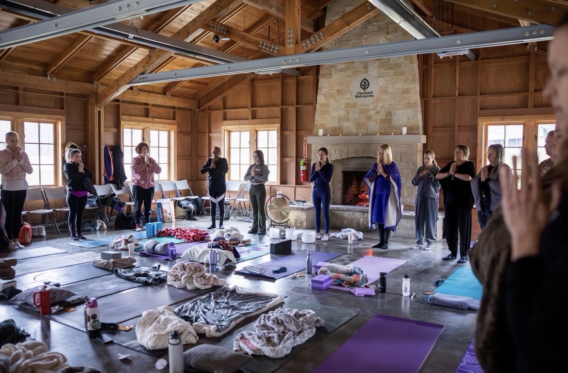 People stand in a circle inside a wooden room with large windows, participating in a group activity with mats and belongings on the floor.