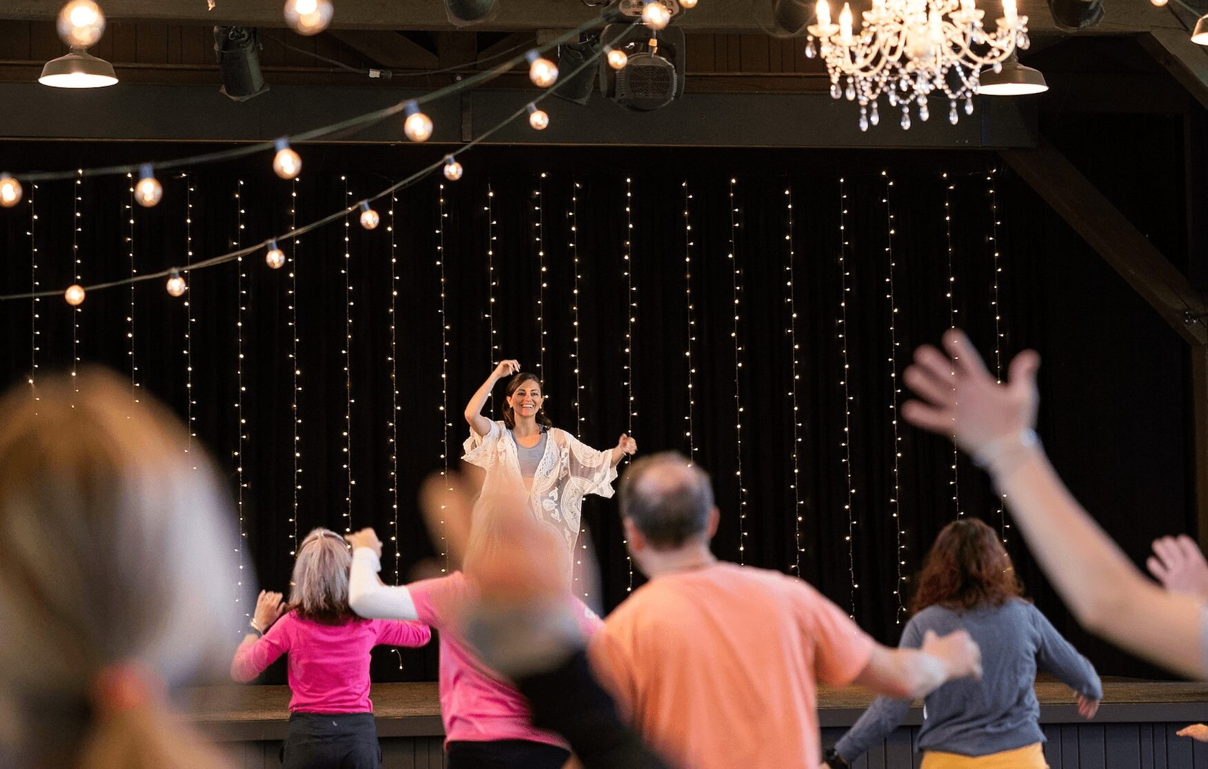 People dancing in front of a stage with a performer, string lights, and a chandelier, in a decorated indoor venue.