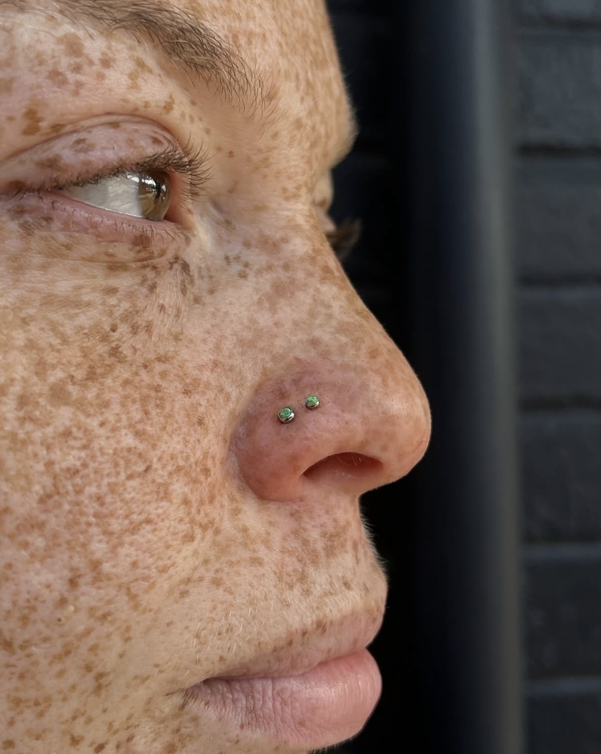 Close-up of a person's face showing freckles, eye, and nose with two nose piercings, against a dark background.