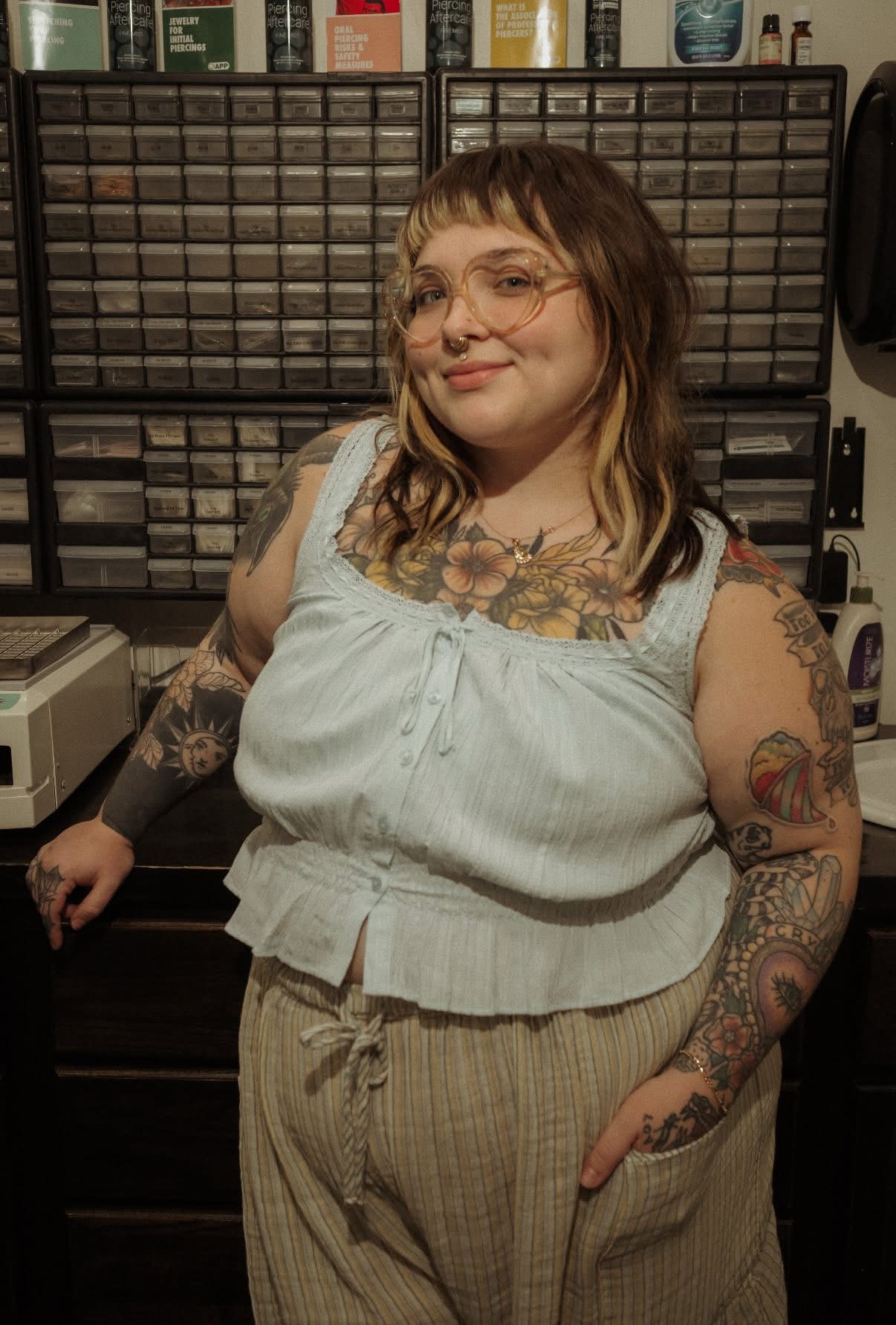 Young woman with glasses and tattoos, wearing a light blue top and beige pants, standing in front of a storage cabinet.