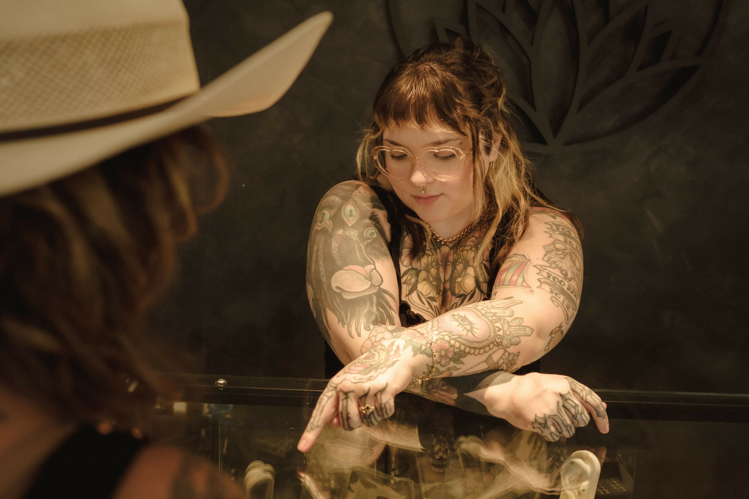 Young woman with tattoos leaning on a glass surface, looking down, with a person in a hat in the foreground.