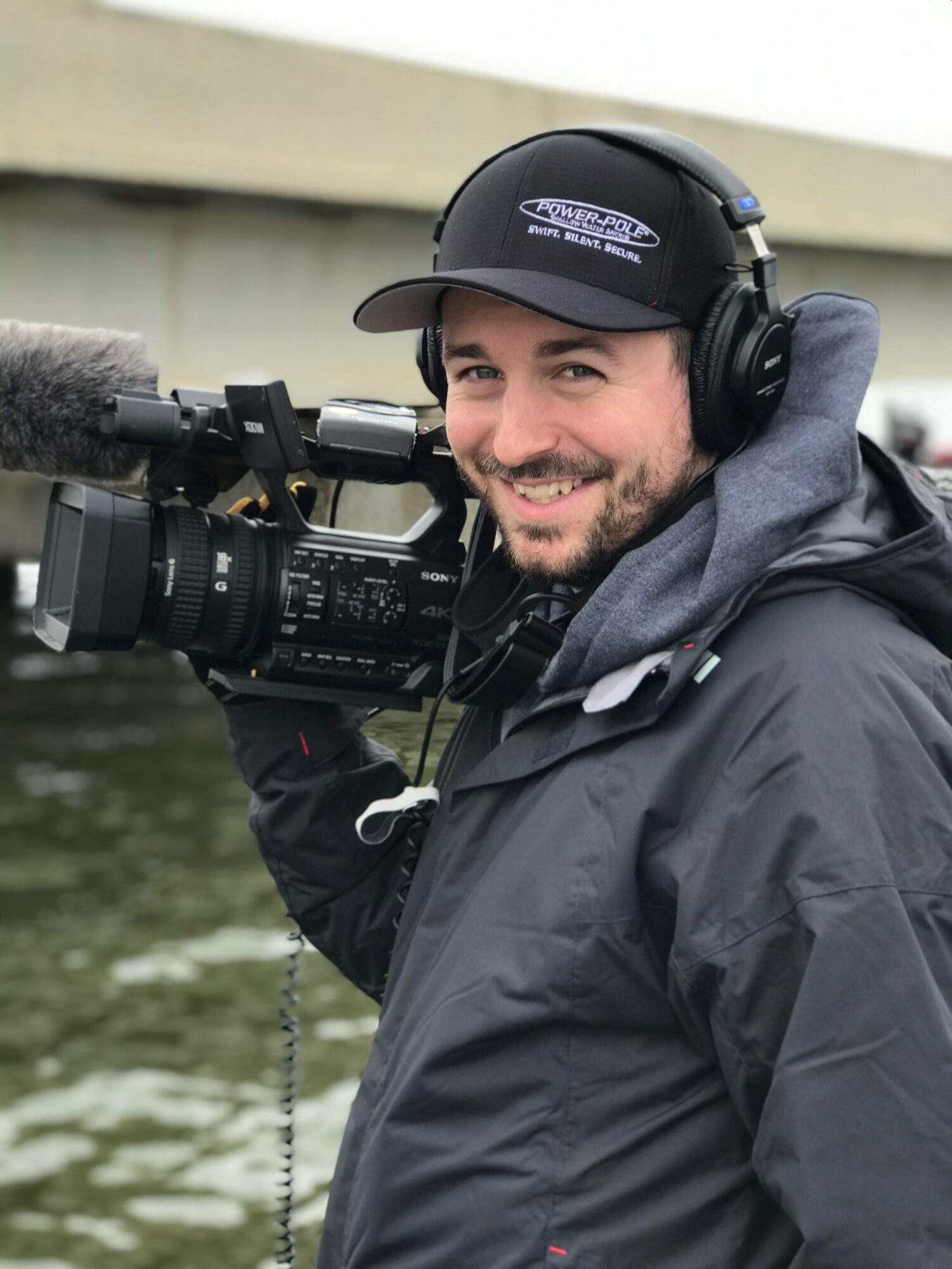 Man with a beard wearing a cap and headphones, holding a professional video camera outdoors near water.