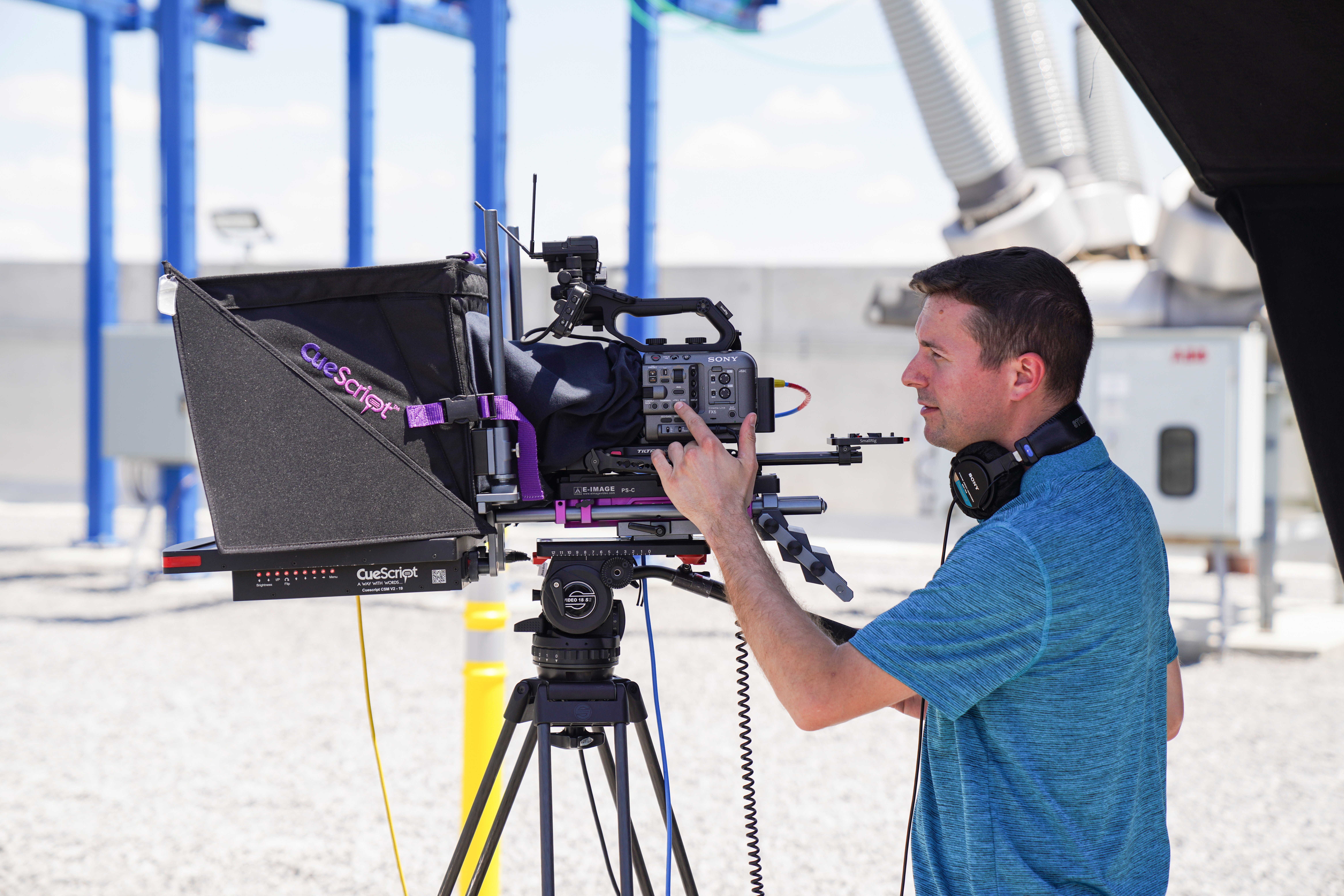 A man operating a professional video camera outdoors with industrial structures in the background.