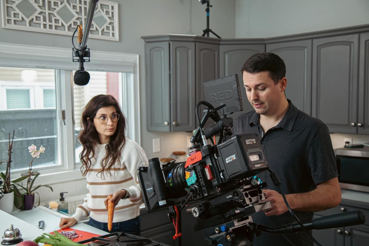 A man and a woman in a kitchen with gray cabinets and a window, preparing to film with professional camera equipment.