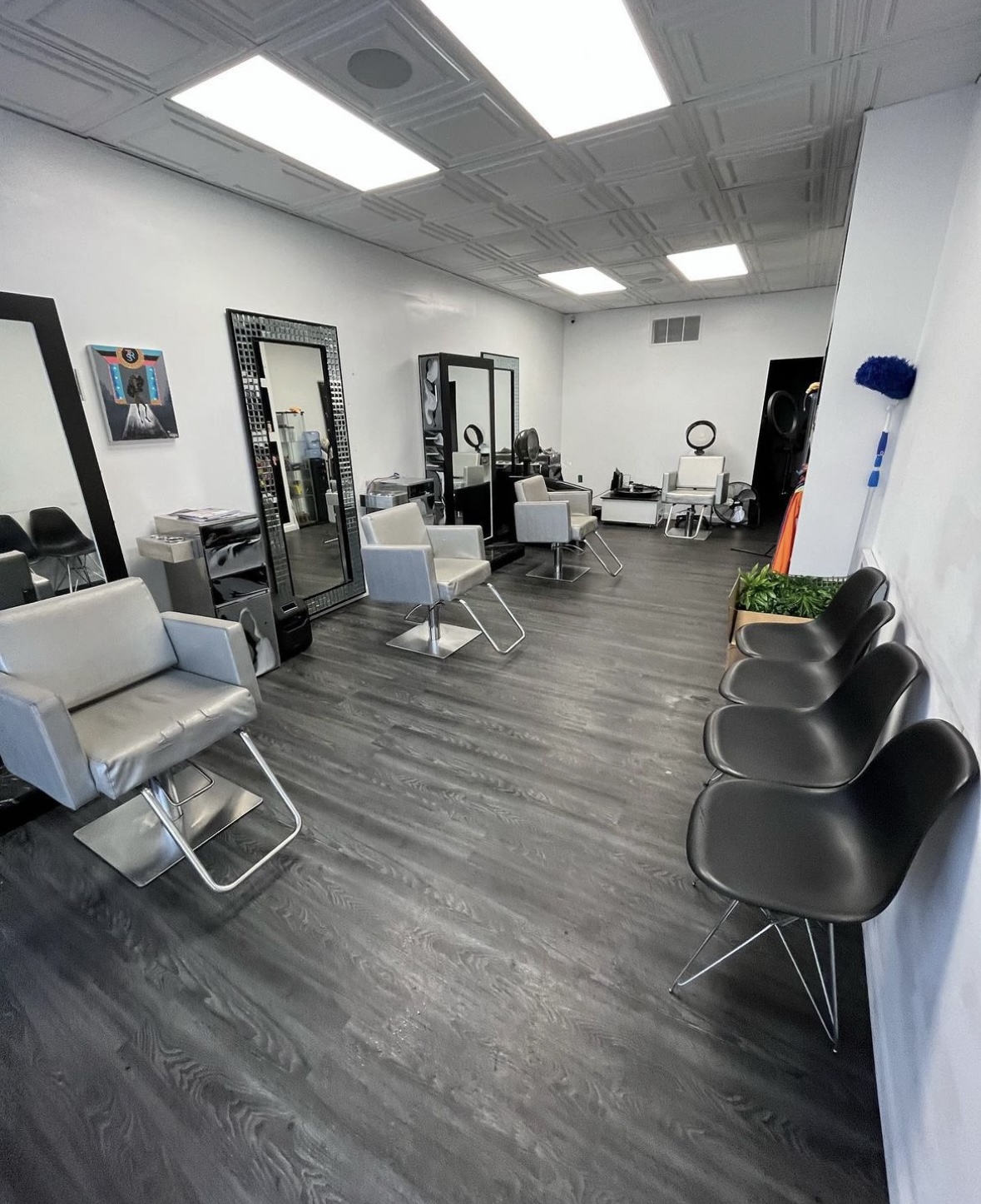 Salon with chairs, mirrors, and styling stations, white walls, and ceiling lights, with a row of black chairs along the wall.