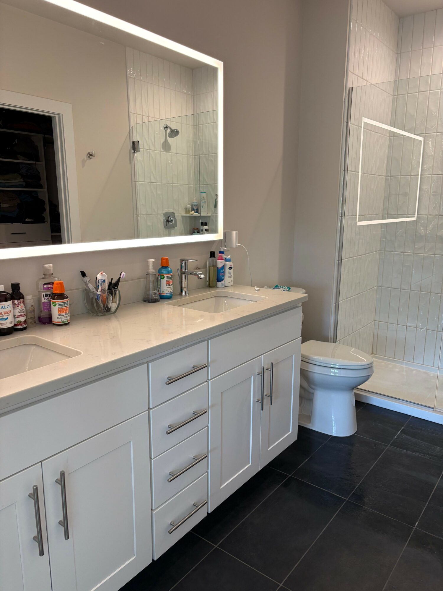 Bathroom vanity with a marble countertop, various toiletries, a white cabinet, and a shower area with a glass door.