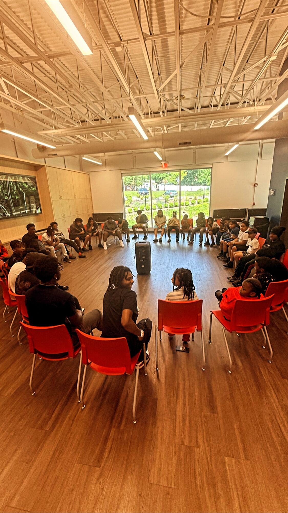 Group of people sitting in a circle in a room with large windows and wooden floors.