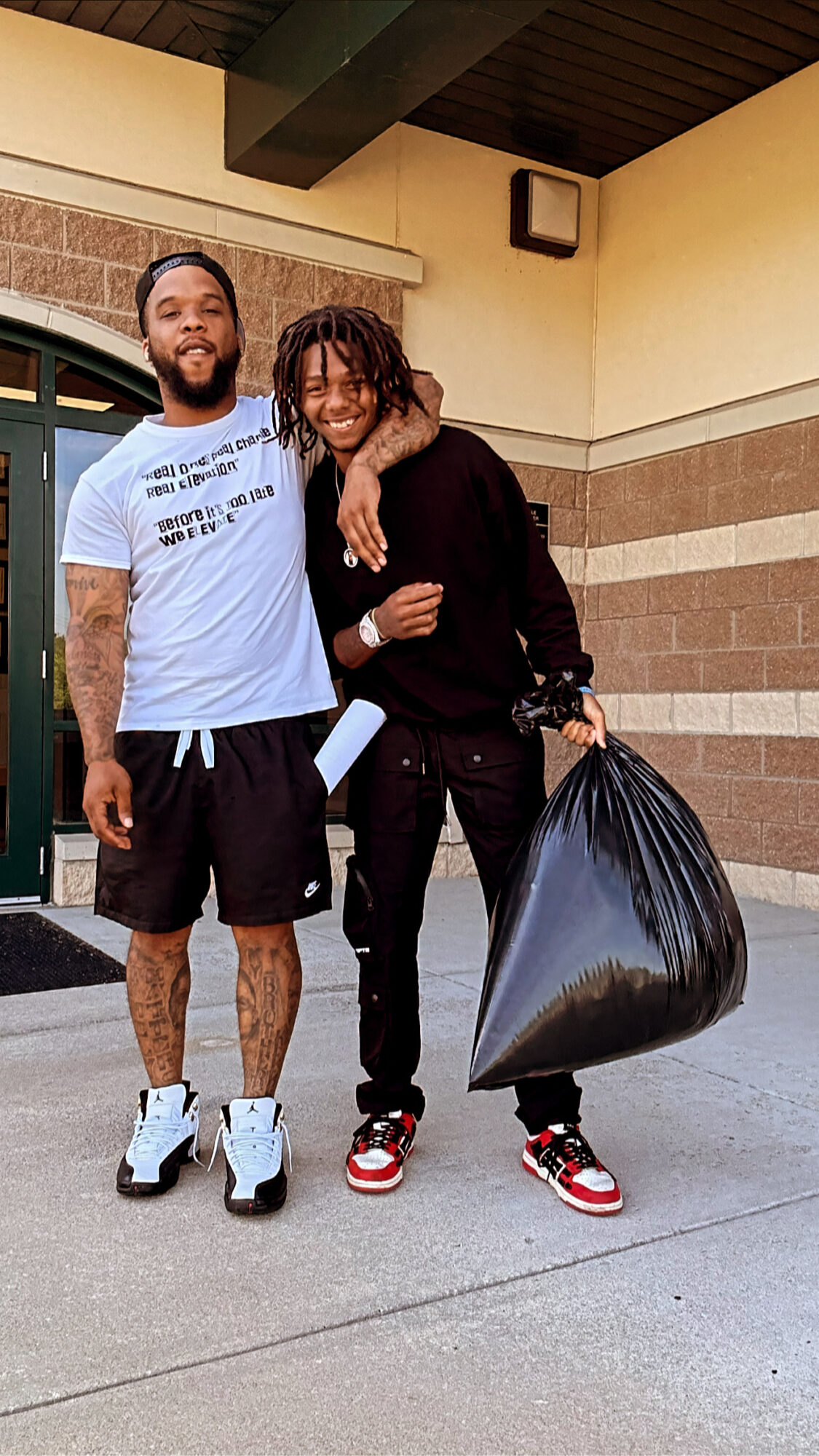 Two men standing outside, one with arm around the other, holding a large trash bag, smiling at the camera.