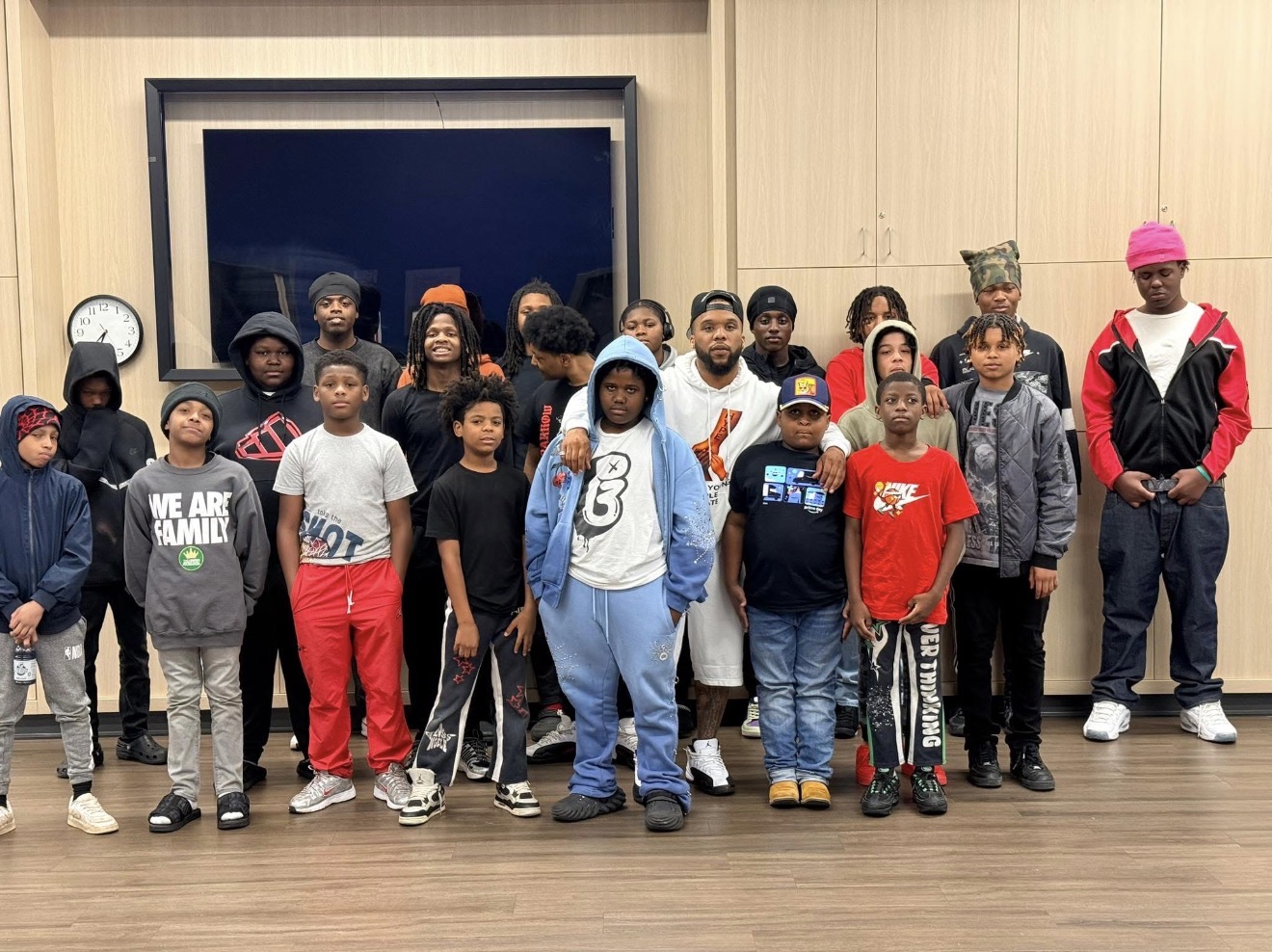 Group of children and adults standing indoors in front of a large window, some wearing colorful clothing and hats.