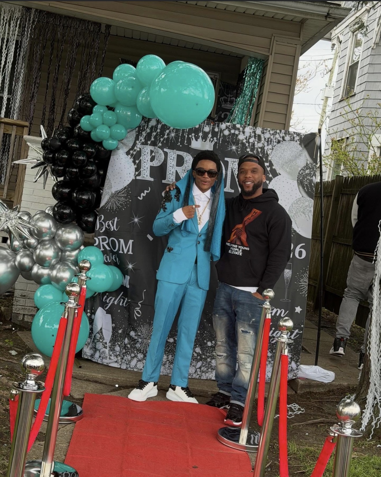 Two people standing on a red carpet in front of a decorated backdrop with balloons, one in a blue suit and sunglasses, the other in casual clothing.