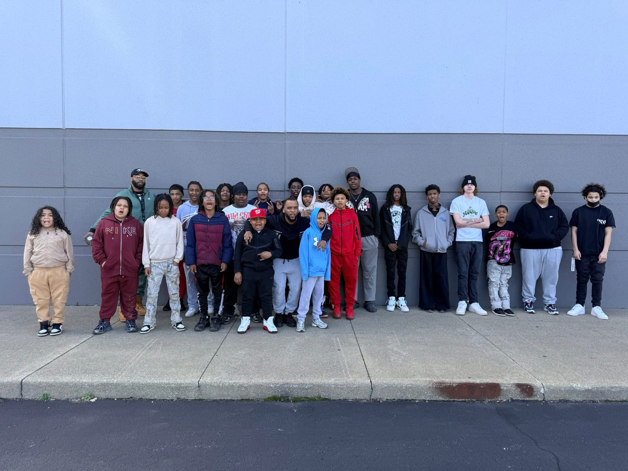 Group of people standing outdoors against a wall, some smiling, wearing casual clothing, with a sidewalk in front.