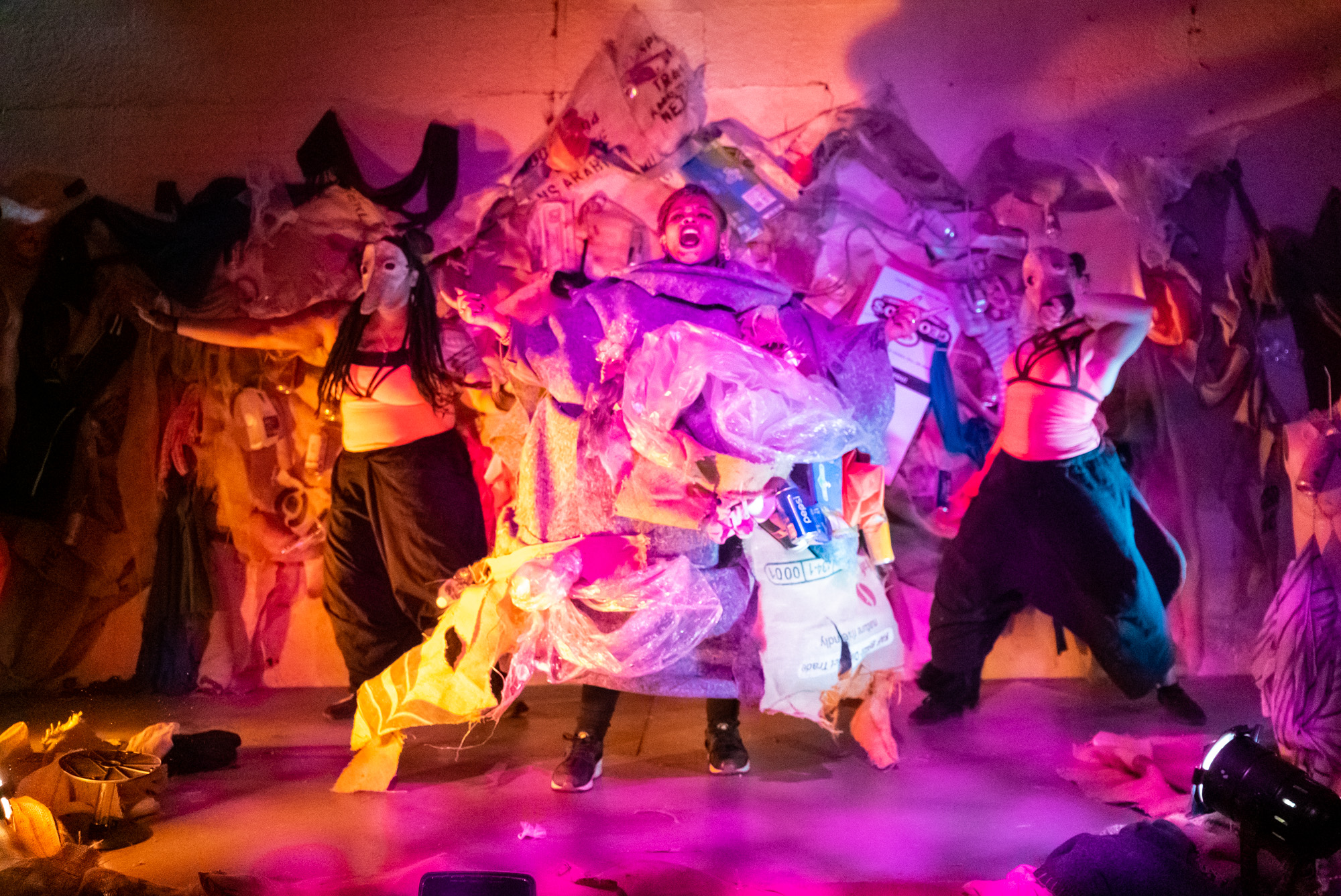 Group of performers dancing on stage with colorful costumes and lighting, surrounded by fabric and props.