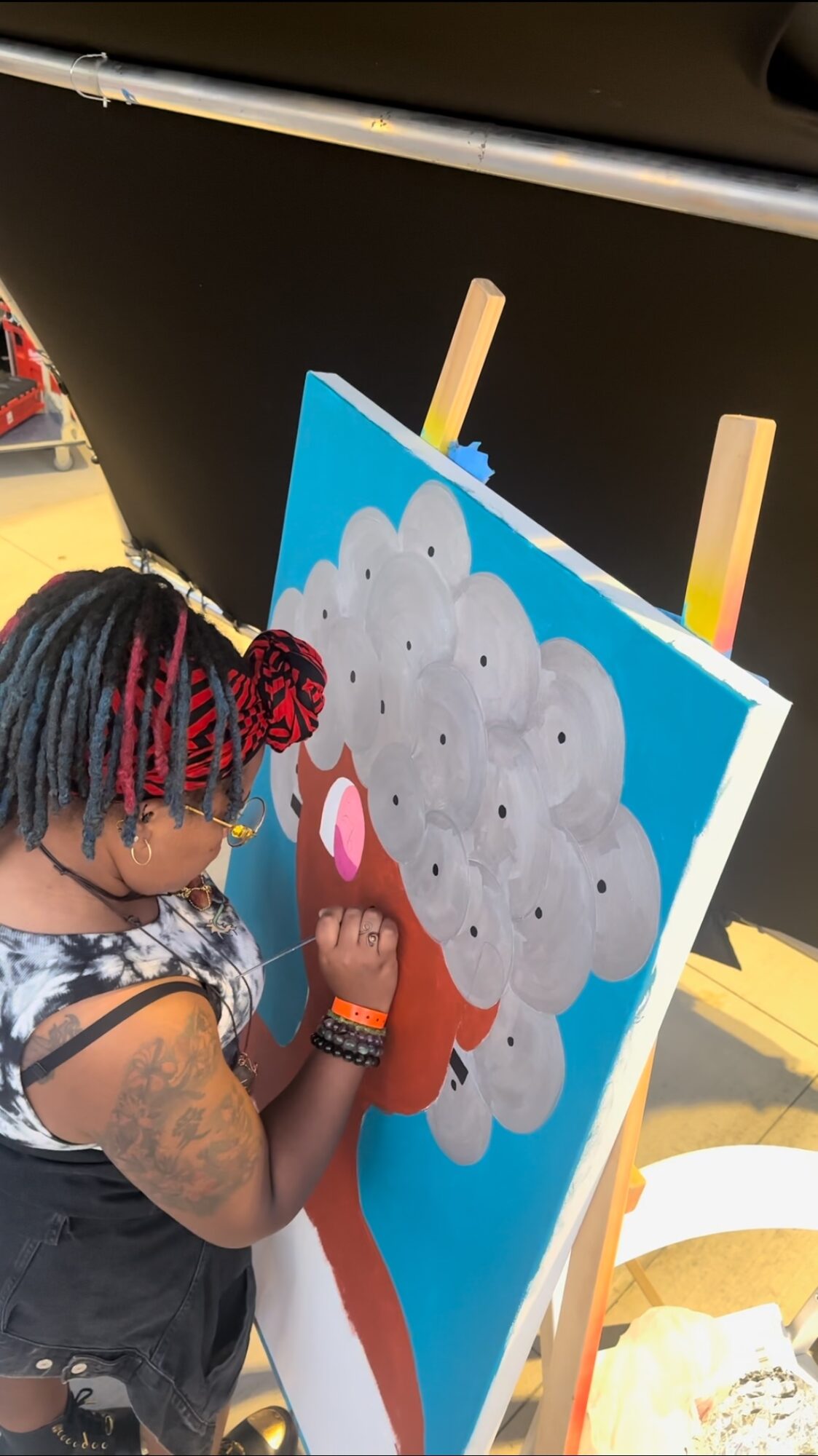 Child painting a cloud with a face on a colorful outdoor easel, with a yellow background and a person with dreadlocks nearby.