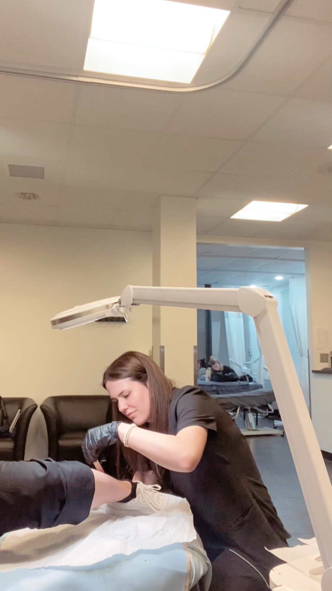 Woman leaning over a table, interacting with a person whose arm is visible, in a room with ceiling lights and a window.