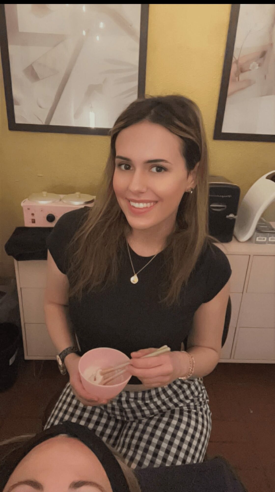 Young woman with long brown hair smiling, holding a pink bowl, sitting in a room with framed pictures on the wall.