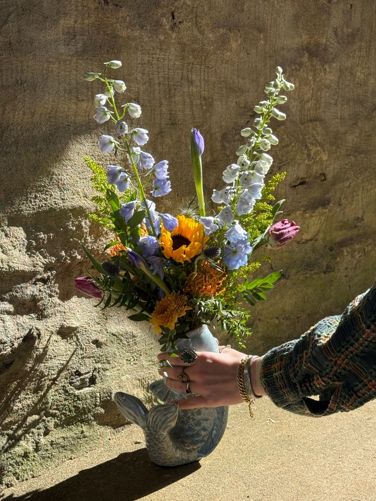 Person holding a colorful flower bouquet in a vase against a textured wall background.