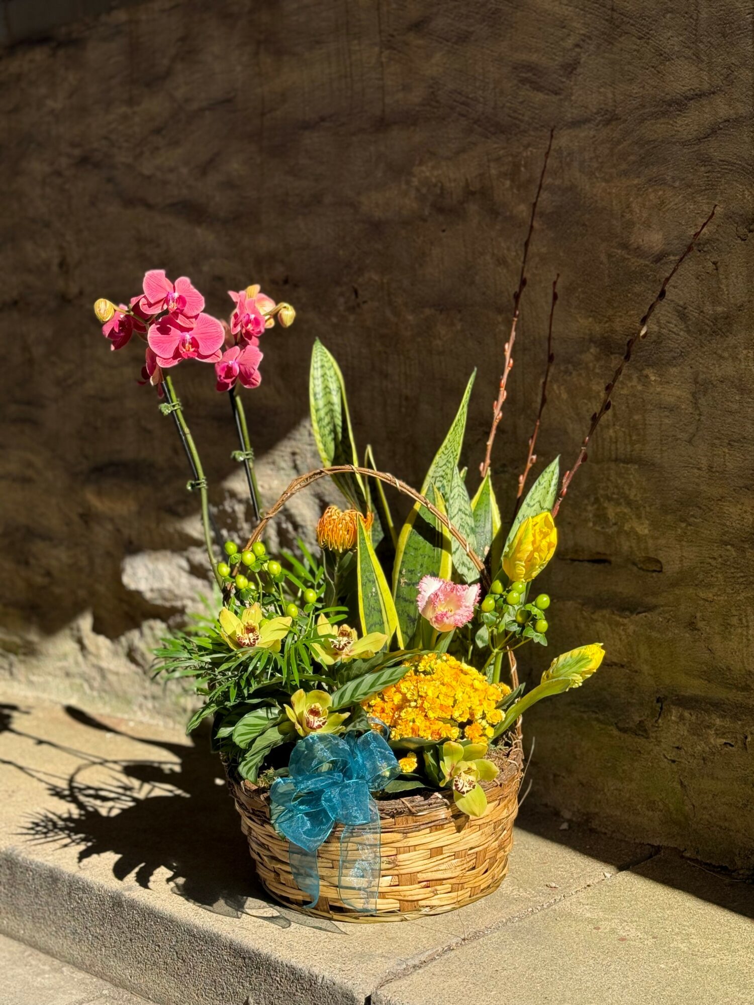 Flower arrangement in a basket with pink, yellow, and green flowers and foliage, placed on a surface against a textured wall.
