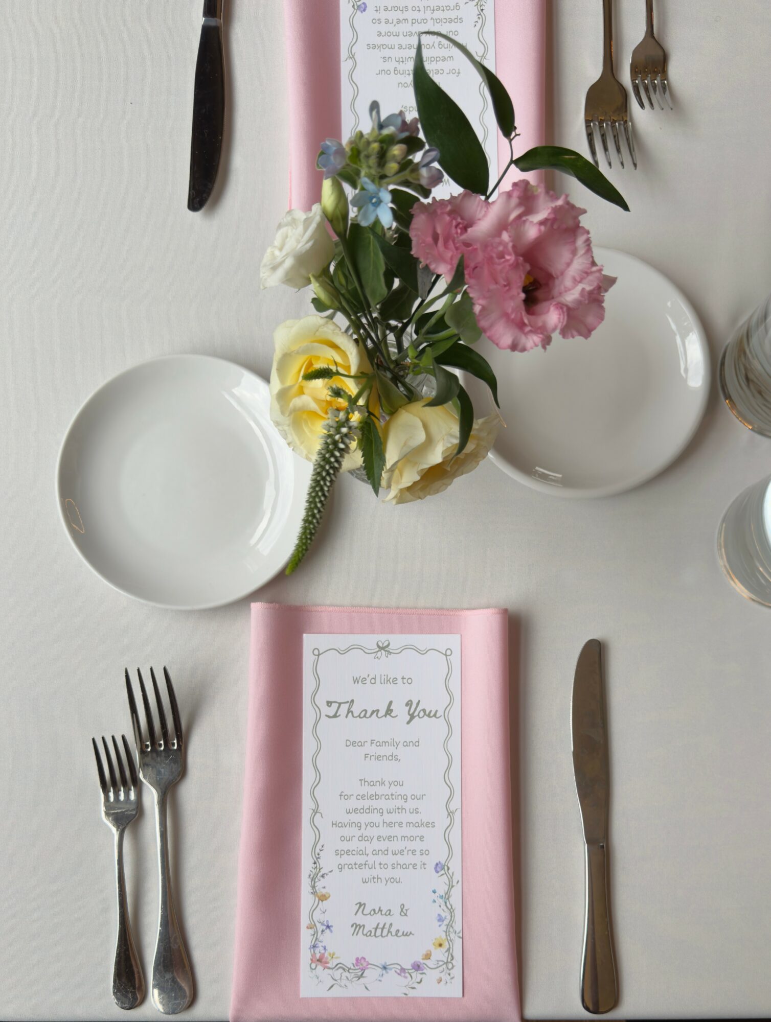 Table setting with a pink napkin, a thank you card, two forks, a knife, a plate, and a flower arrangement.