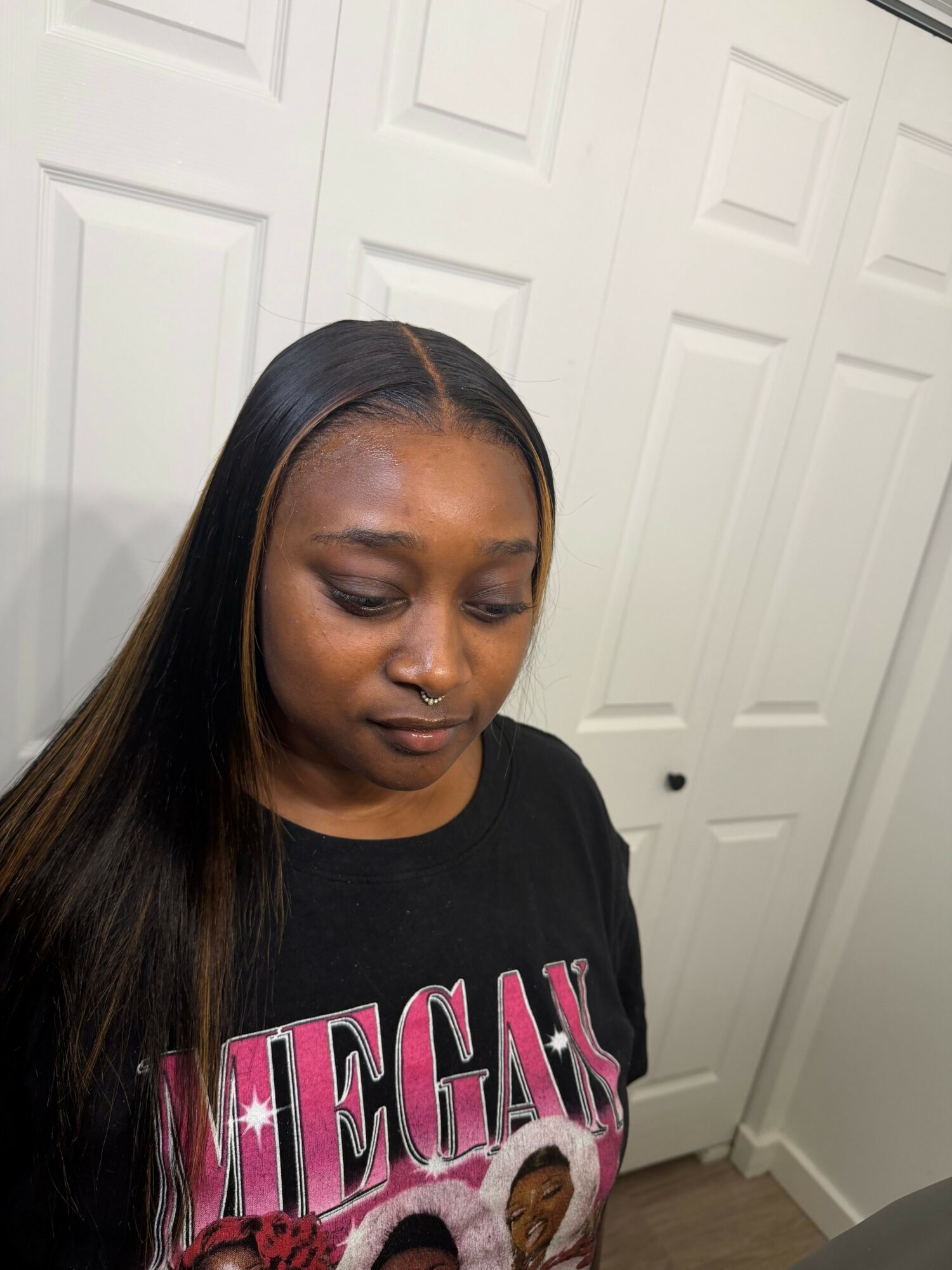 Woman with long straight hair and makeup, wearing a black T-shirt with pink and white text, standing indoors.
