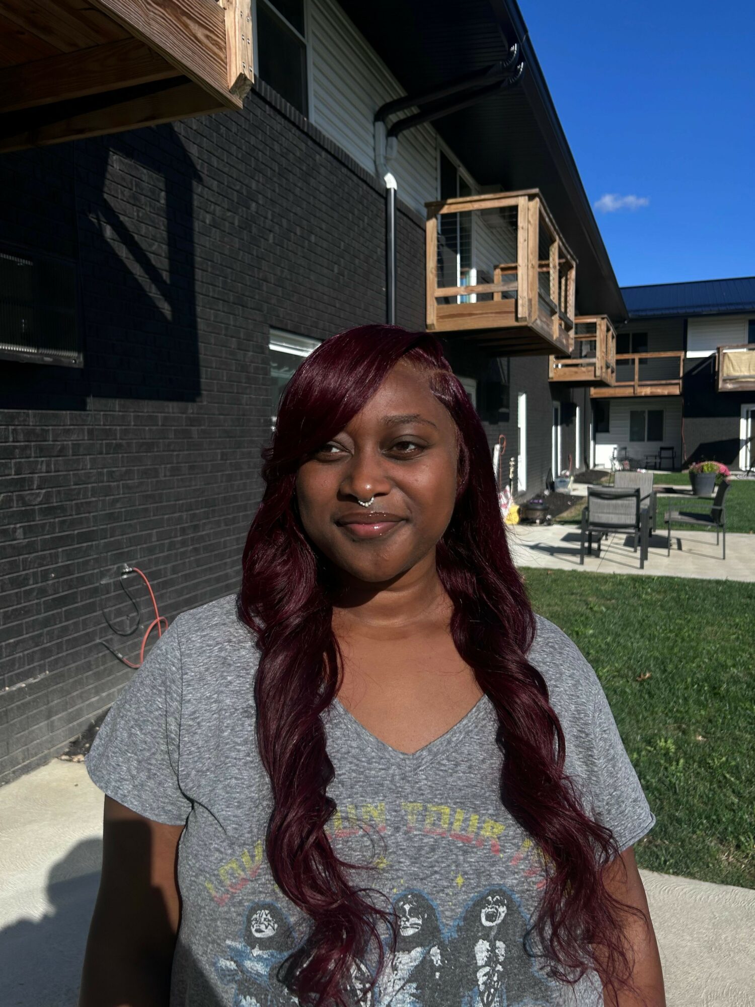 Woman with long, wavy hair smiling outdoors on a sunny day, with buildings and chairs in the background.