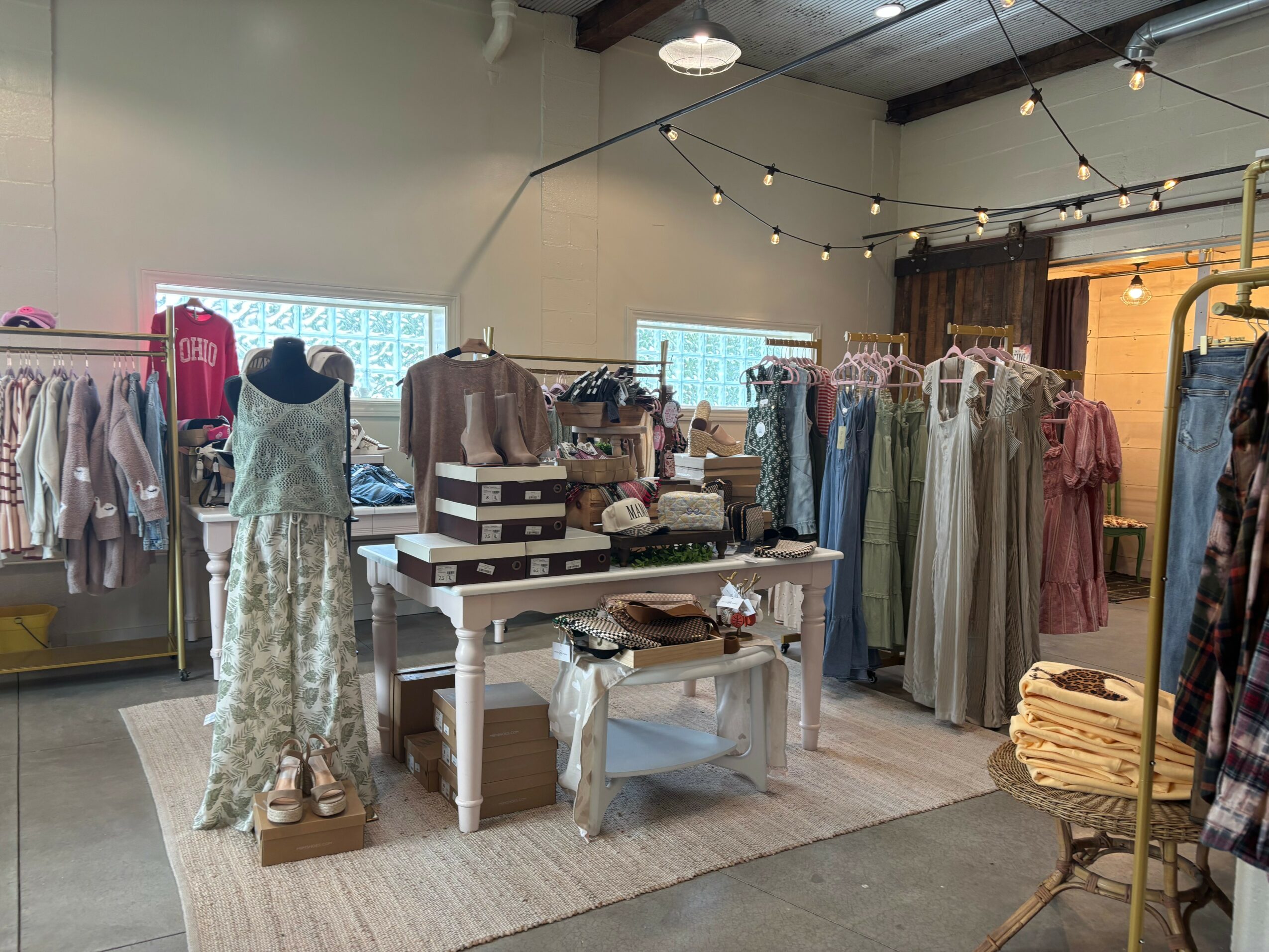 Clothing store interior with dresses, mannequins, and display tables, decorated with string lights and wooden accents.