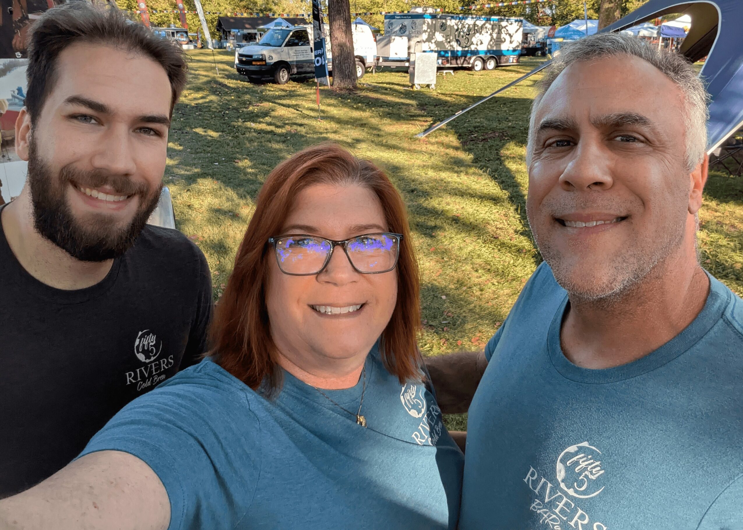 Three people smiling outdoors, two men and one woman, with trees and vehicles in the background.