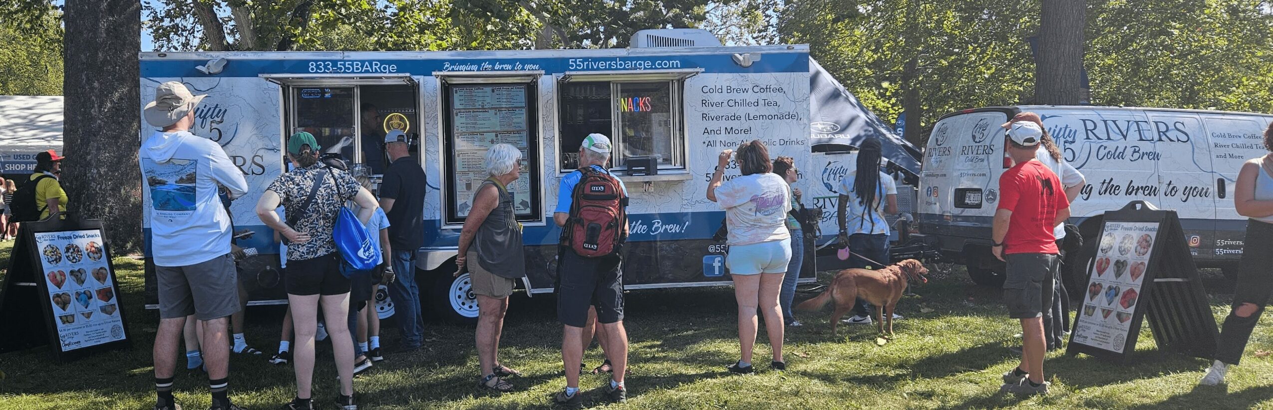 People line up at an ice cream truck outdoors on a sunny day, with trees and parked vehicles in the background.