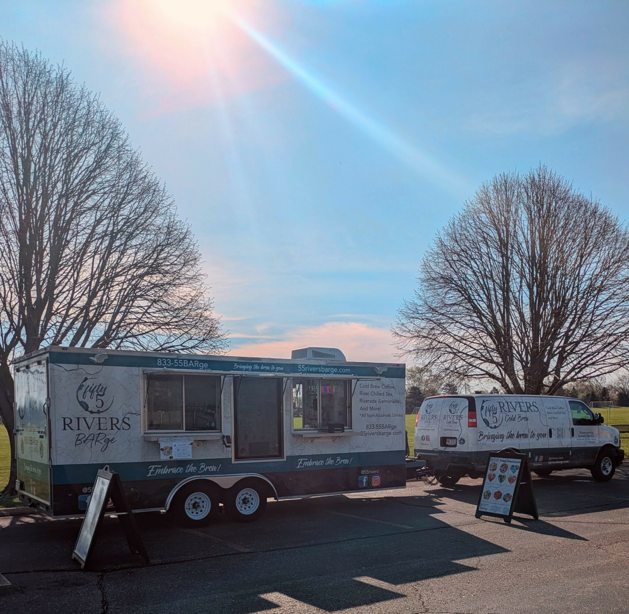 Food truck and van parked outdoors with leafless trees and a clear sky, sunlight shining brightly, and signs on the ground.