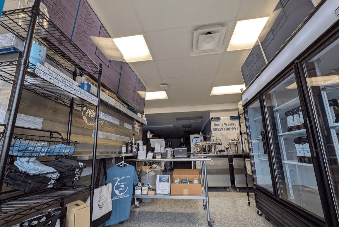 Interior of a store with shelves, a refrigerated display case, and a checkout counter, lit by ceiling lights.