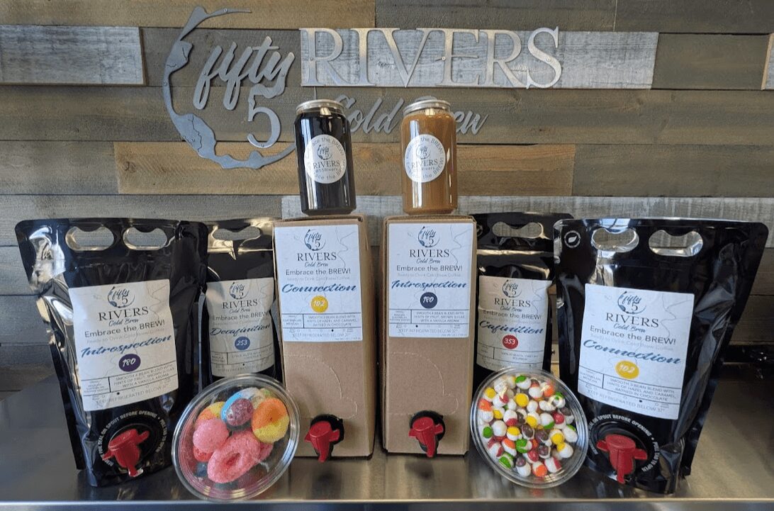 Display of bottled beverages, bags of snacks, and bowls of colorful candies on a wooden surface with a sign in the background.