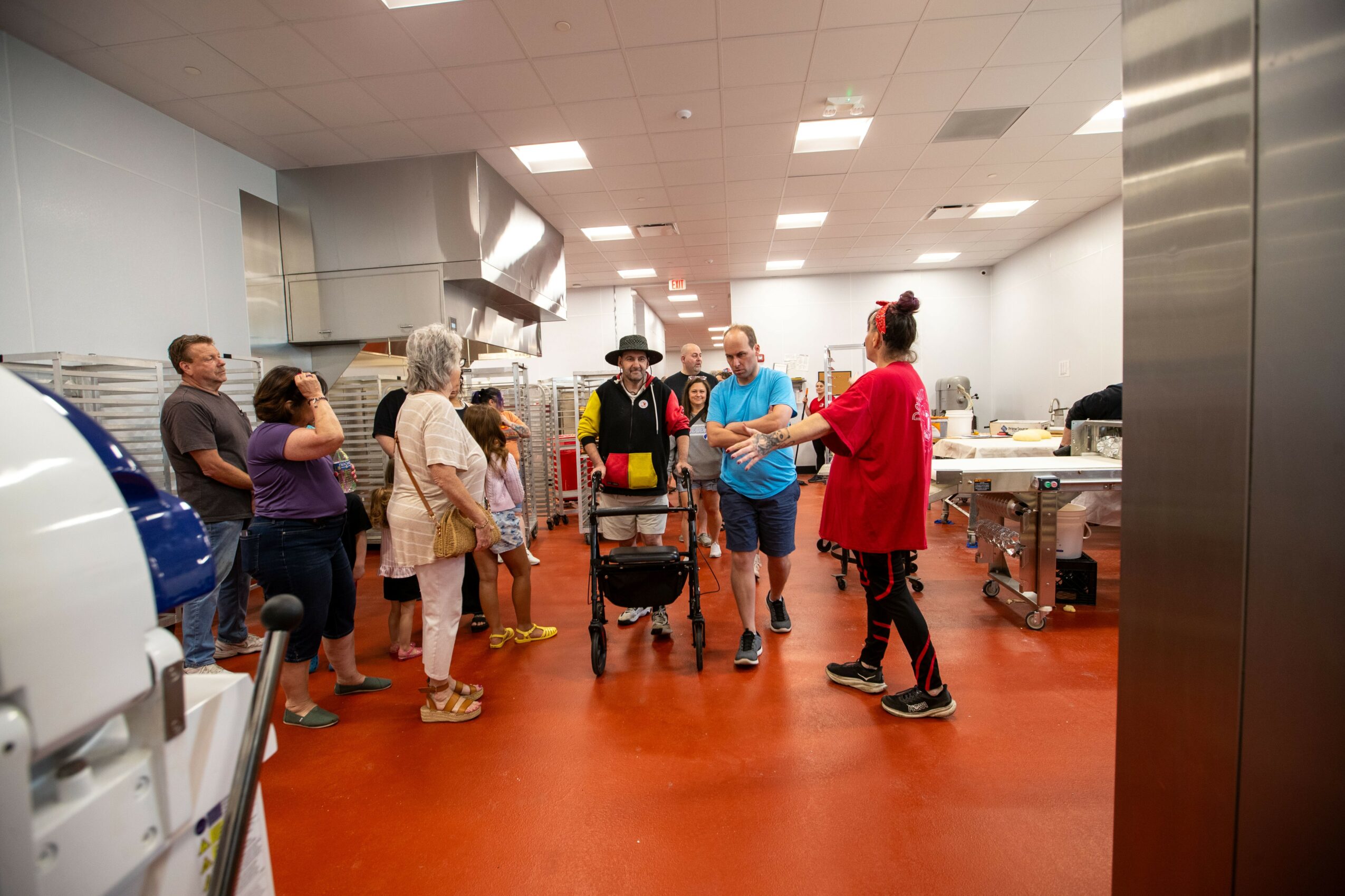 Group of people gathered in a kitchen or cafeteria, listening to a woman in a red jacket speak, with a cart and kitchen equipment visible.