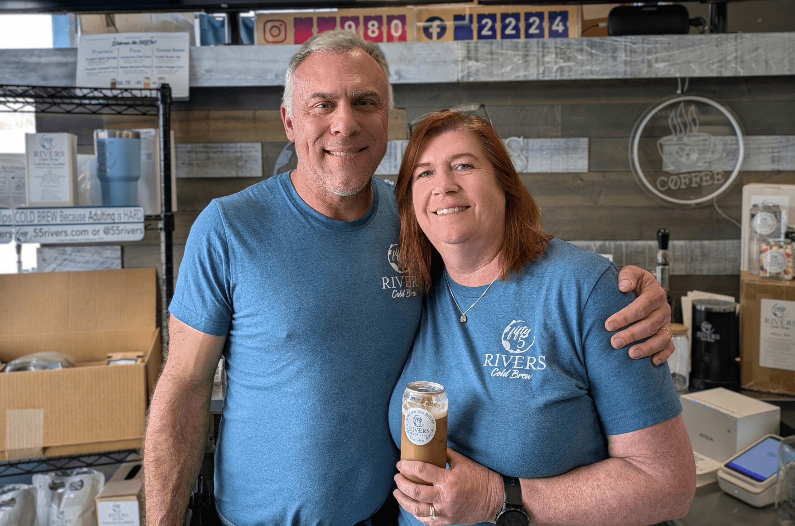 Two smiling people in blue shirts standing together inside a store, holding a can, with shelves and boxes behind them.