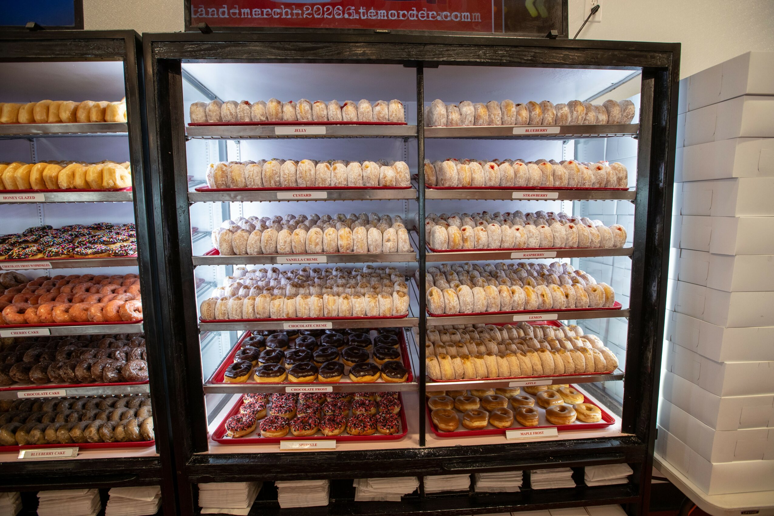 Glass display case filled with assorted cookies and baked goods on multiple shelves, with a white wall and boxes nearby.