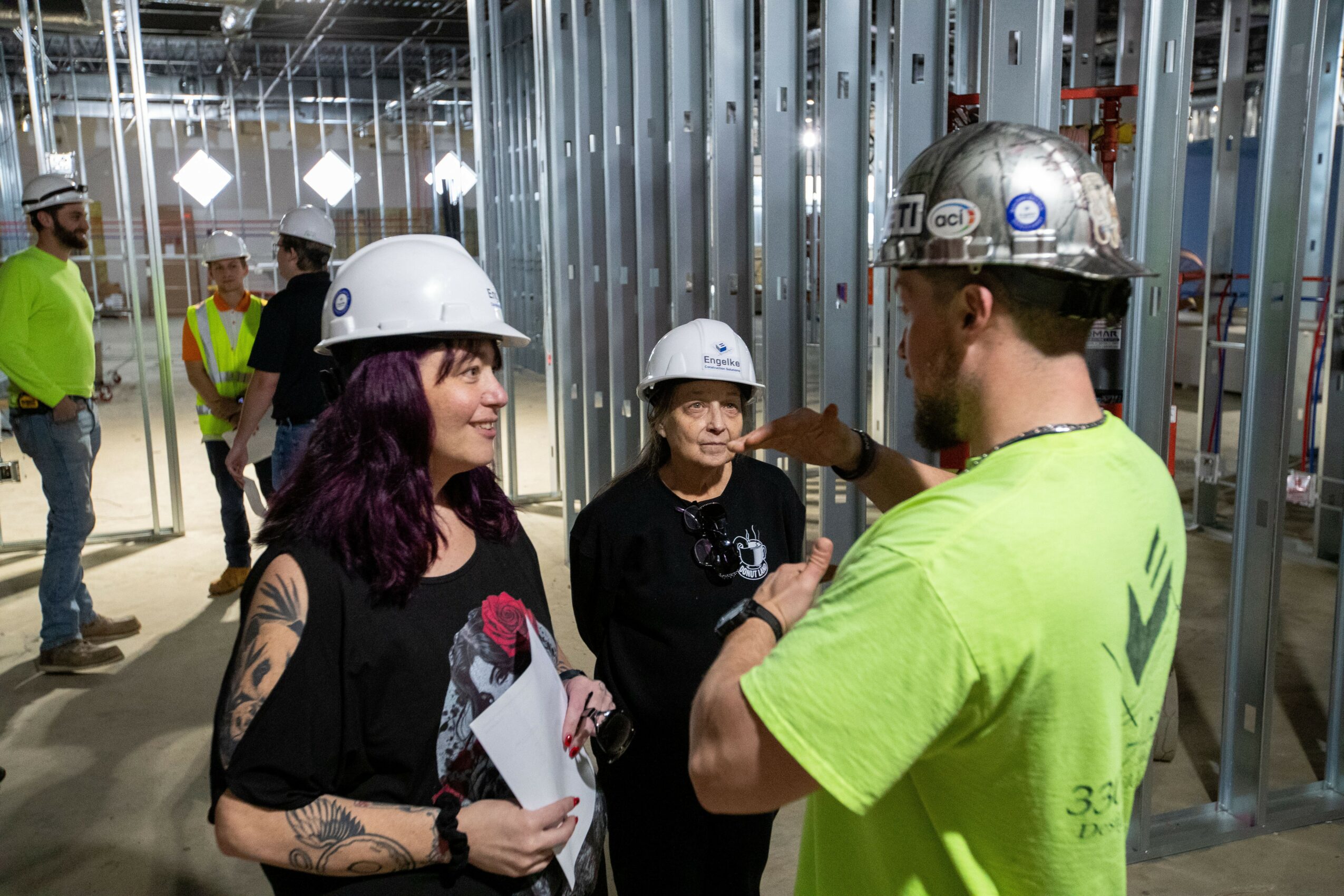 Group of construction workers and a woman in a building under construction, talking and wearing safety helmets and vests.