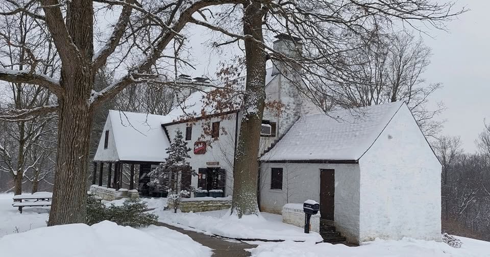 Snow-covered house with trees and a small white building nearby, winter scene, snow on ground and roof, overcast sky.