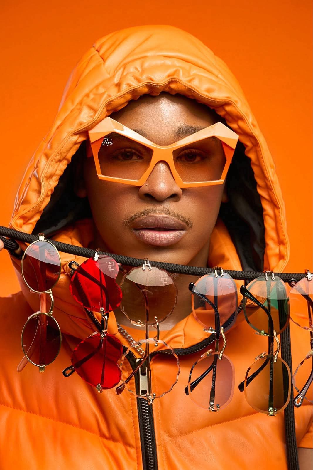 Person wearing orange sunglasses and hooded orange jacket, standing behind a display of sunglasses against an orange background.