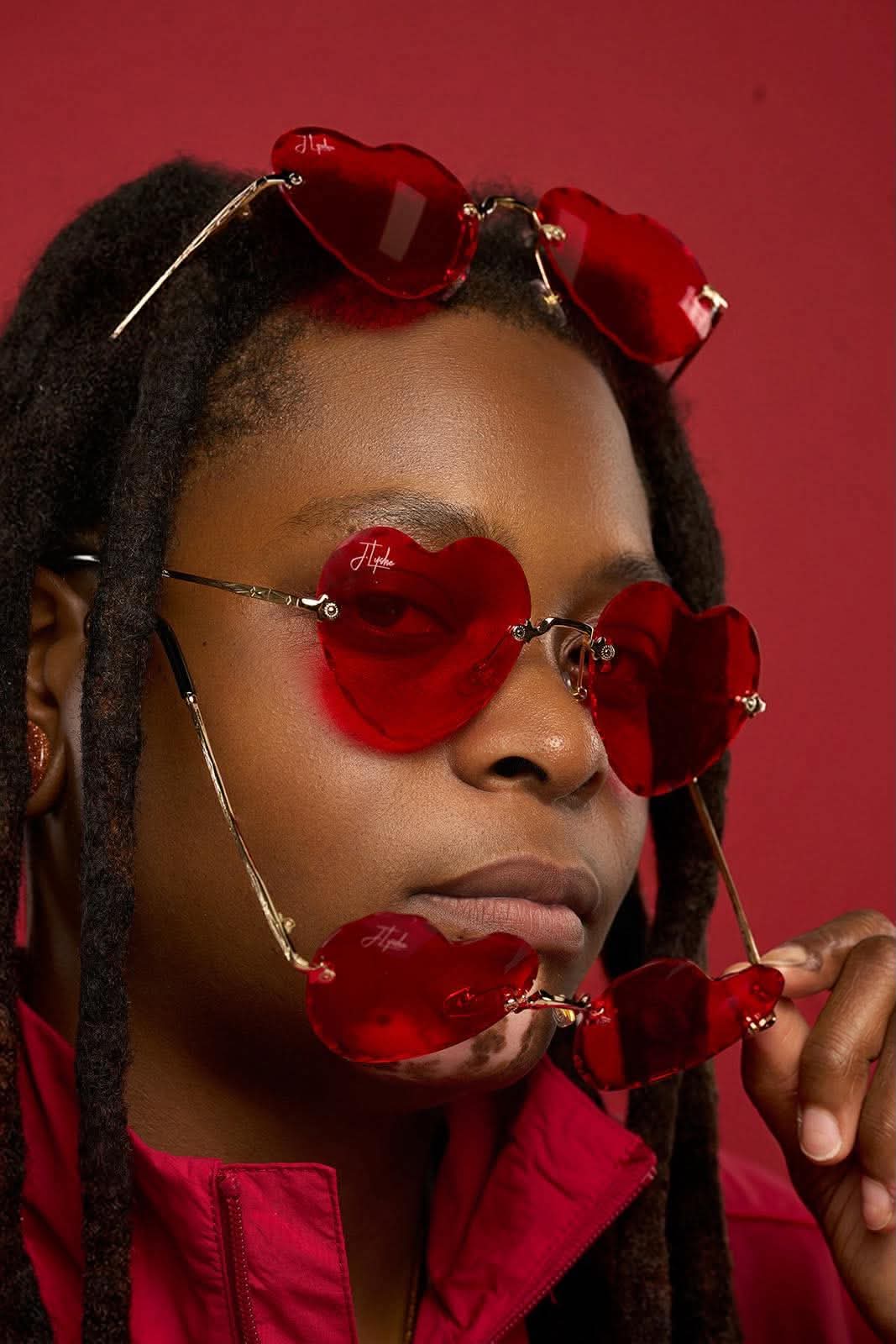 Person with heart-shaped red sunglasses and red accessories, holding a red object near face, against pink background.