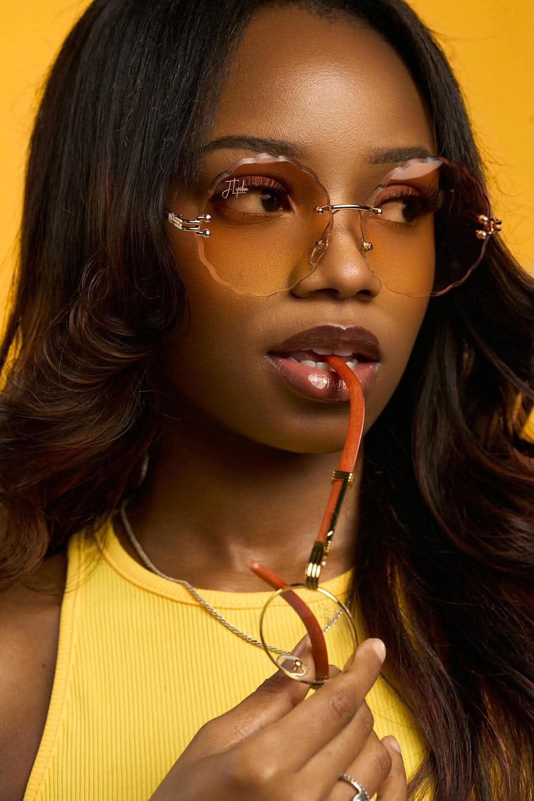 Woman with long dark hair wearing yellow sunglasses and a yellow top, holding glasses near her lips, against a yellow background.