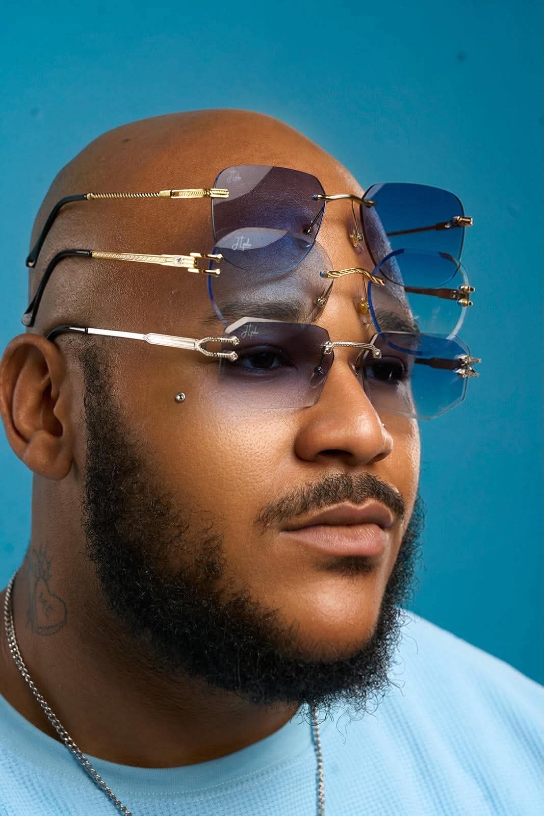 Man with four pairs of sunglasses stacked on his face, bald head, beard, against a blue background.