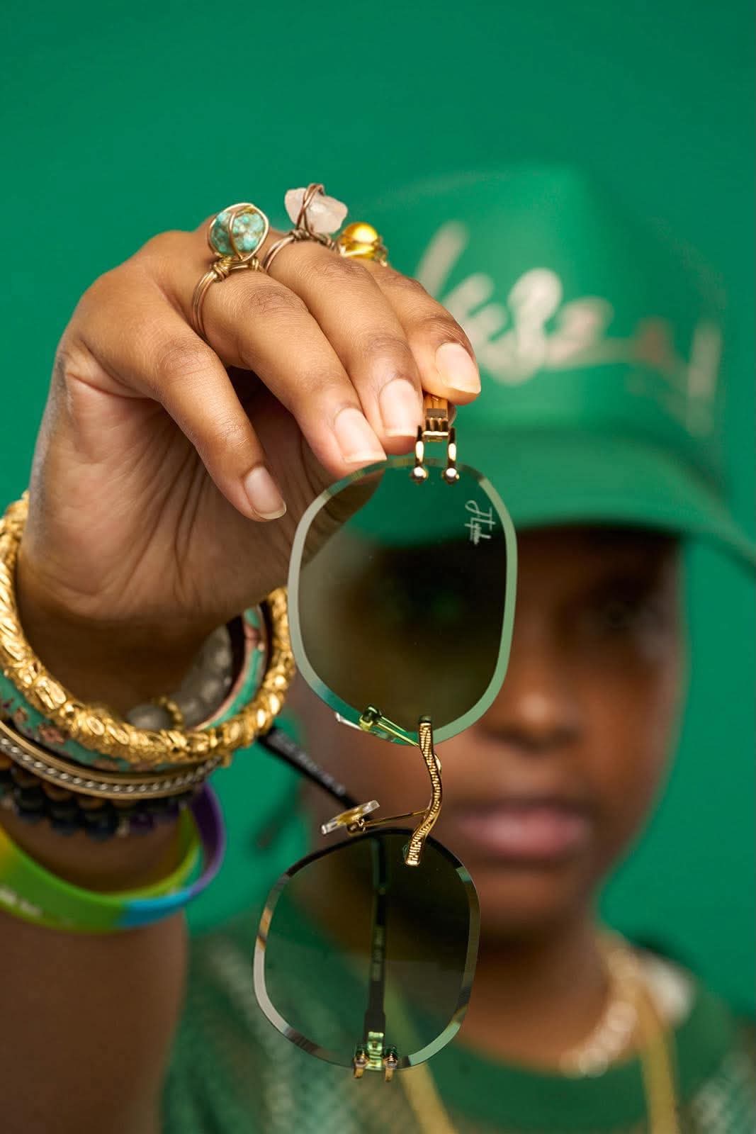 Person holding sunglasses with reflection, wearing multiple rings and bracelets, with a green cap and blurred face in background.