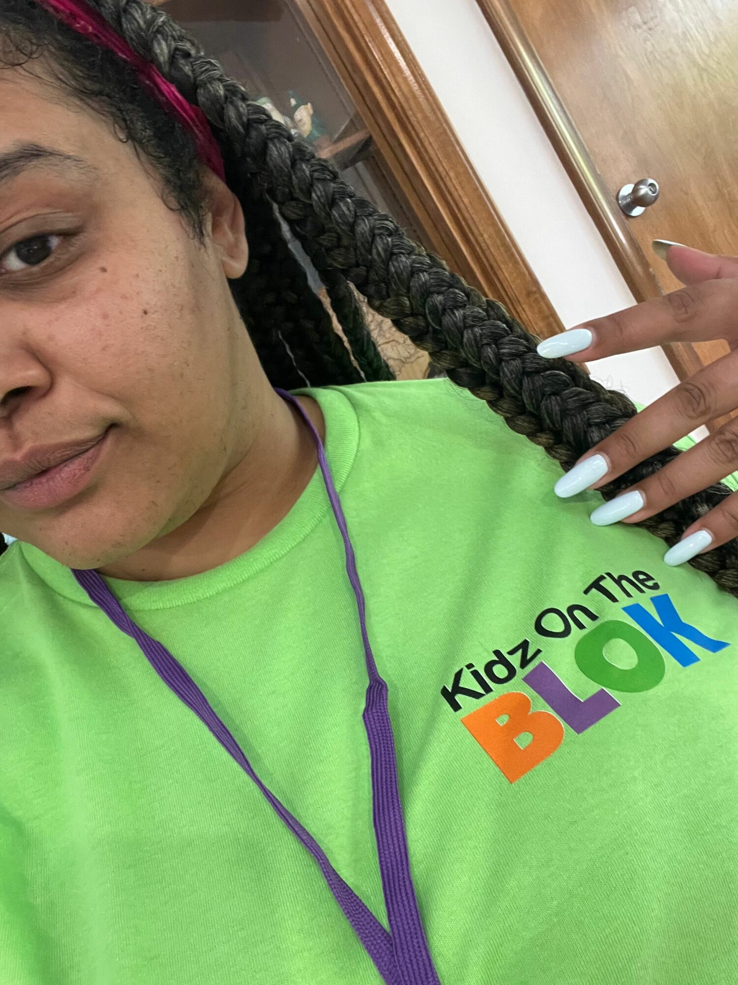Person with long braided hair, wearing a bright green shirt with colorful text, holding braid, with wooden surface in background.