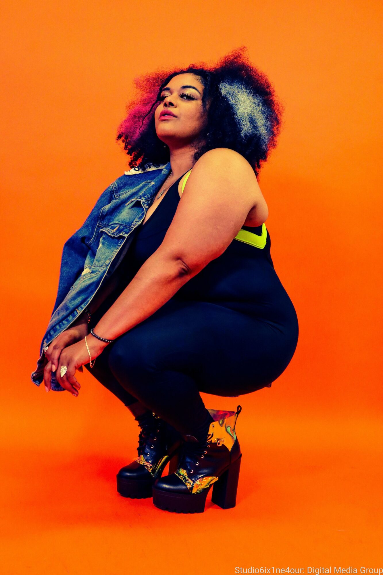Woman with curly hair squatting against an orange background, wearing a denim vest, black outfit, and high-heeled shoes.
