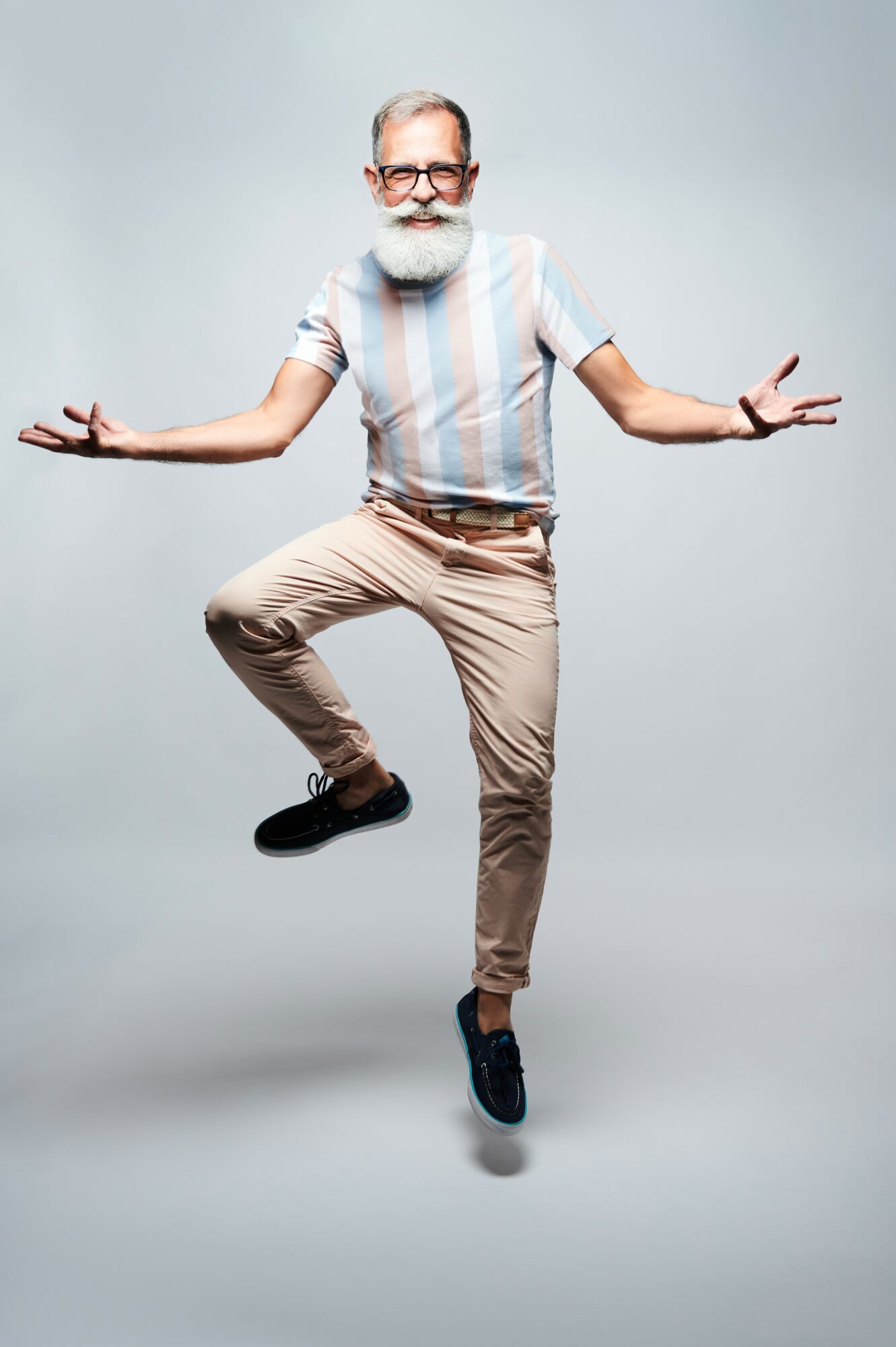 Older man with glasses and a beard balancing on one leg with arms outstretched, smiling, against a plain background.
