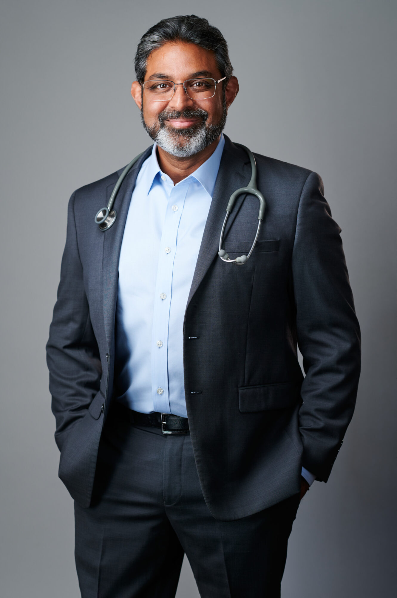 A smiling male doctor with glasses, wearing a suit and stethoscope, standing against a gray background.
