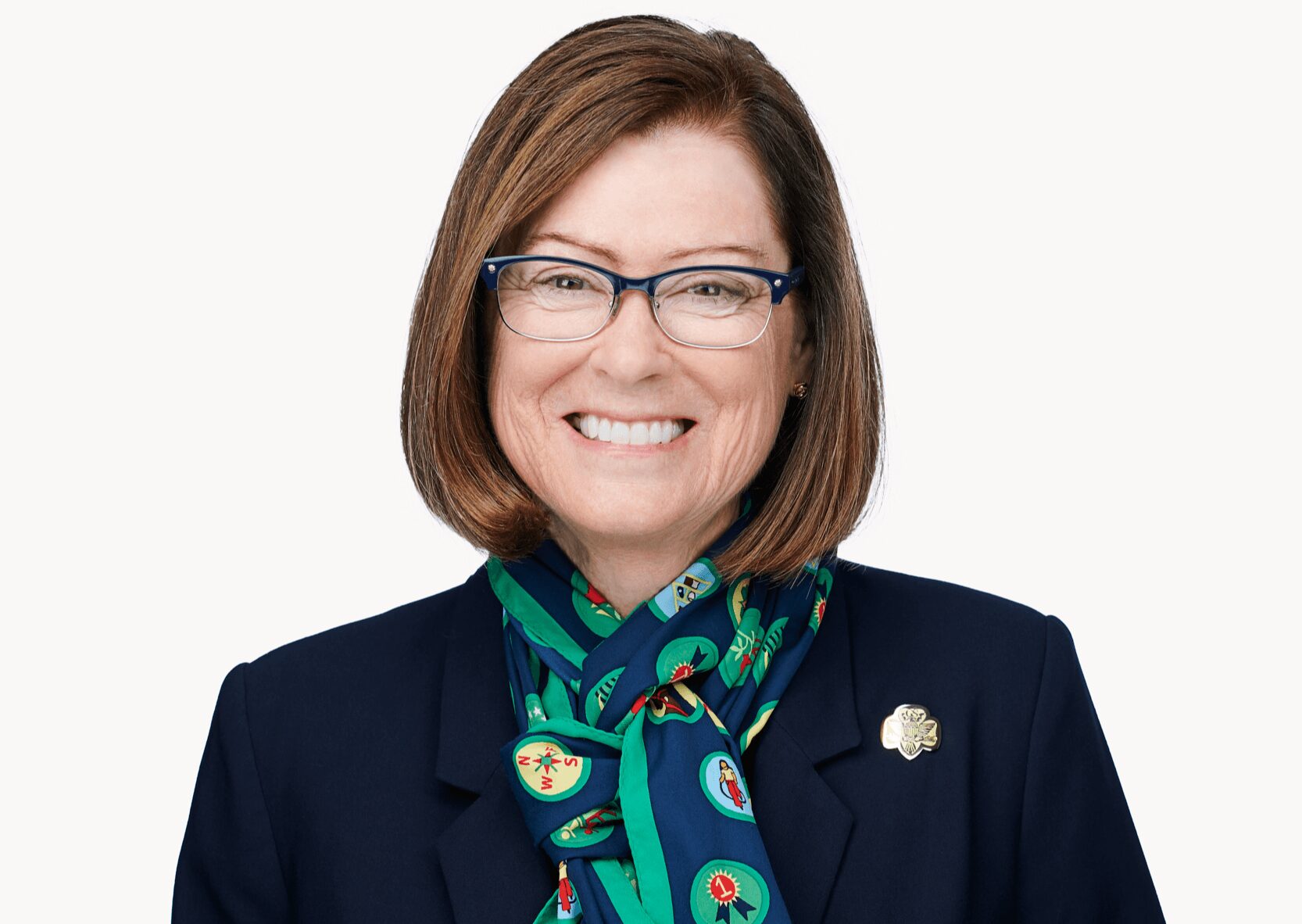 Portrait of a smiling woman with glasses, wearing a dark blazer and a colorful scarf, against a plain background.