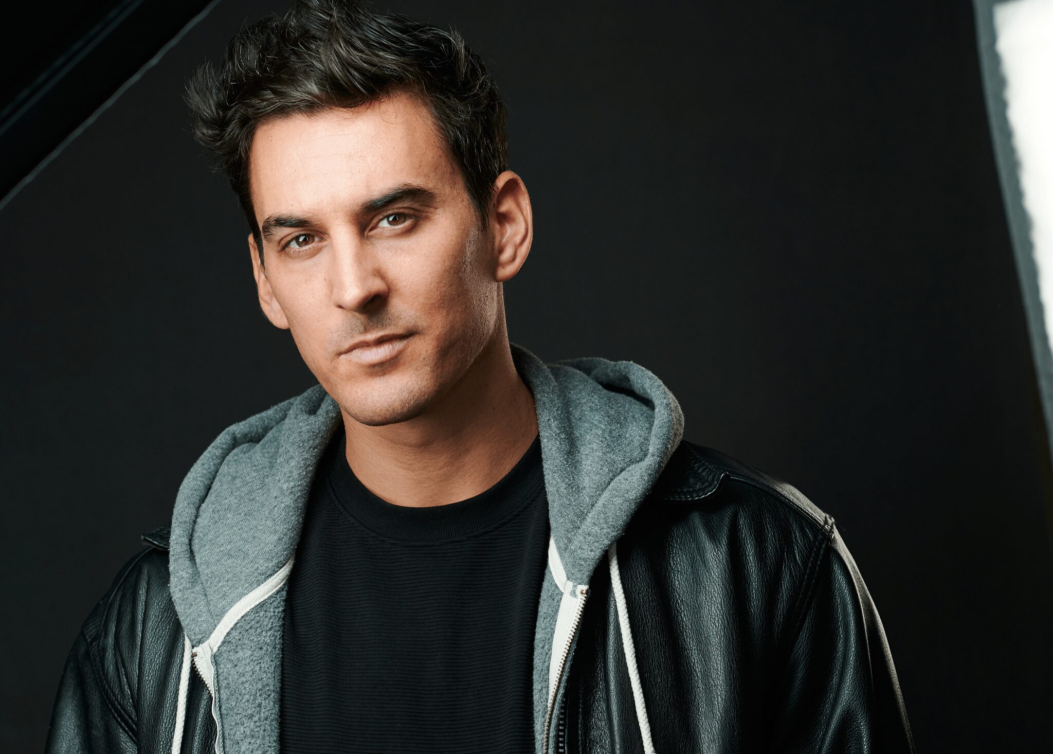 Young man with dark hair, wearing a black leather jacket and gray hoodie, looking at the camera against a dark background.