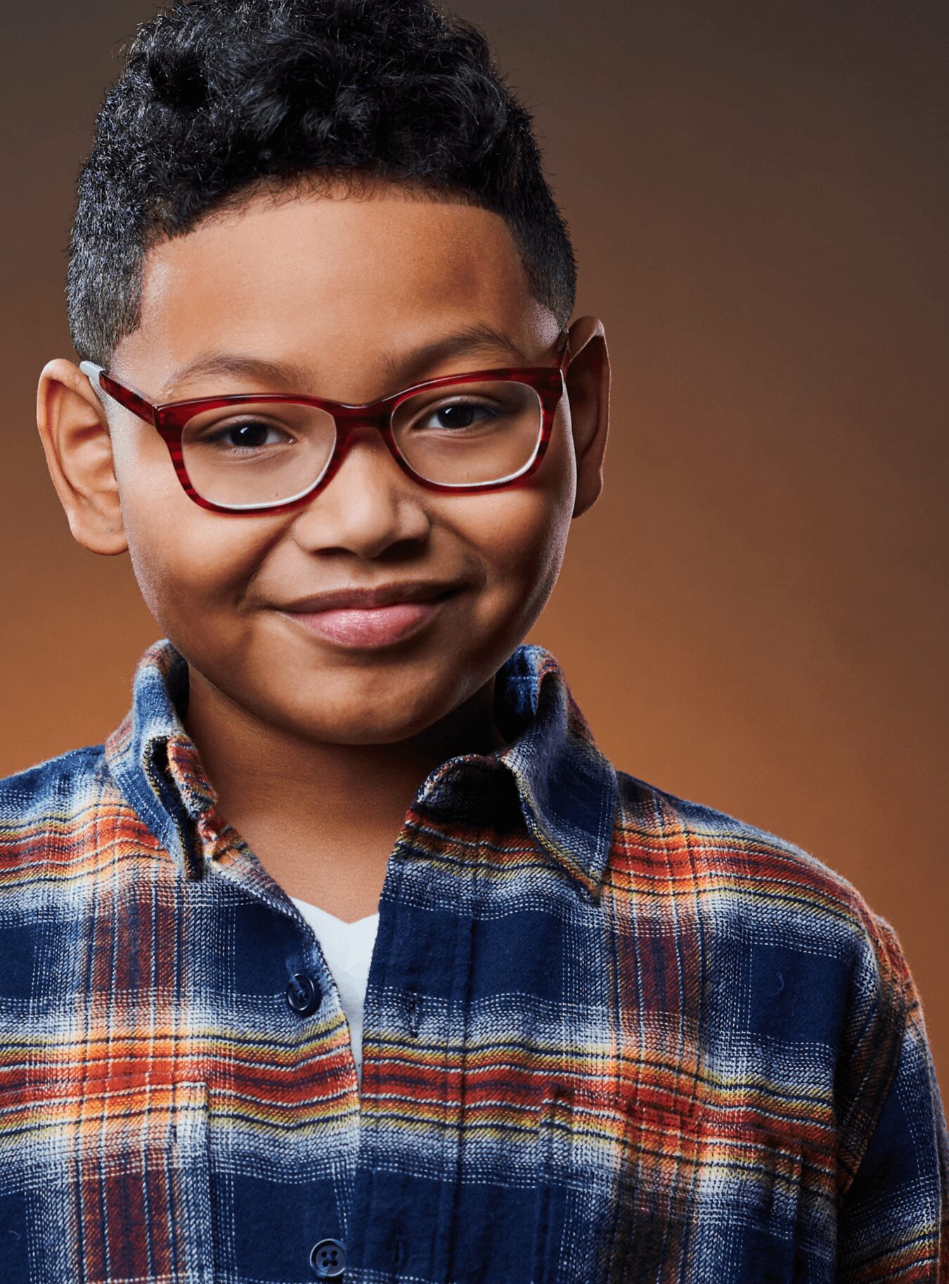Young person with short curly hair, red glasses, and a plaid shirt smiling, facing forward.
