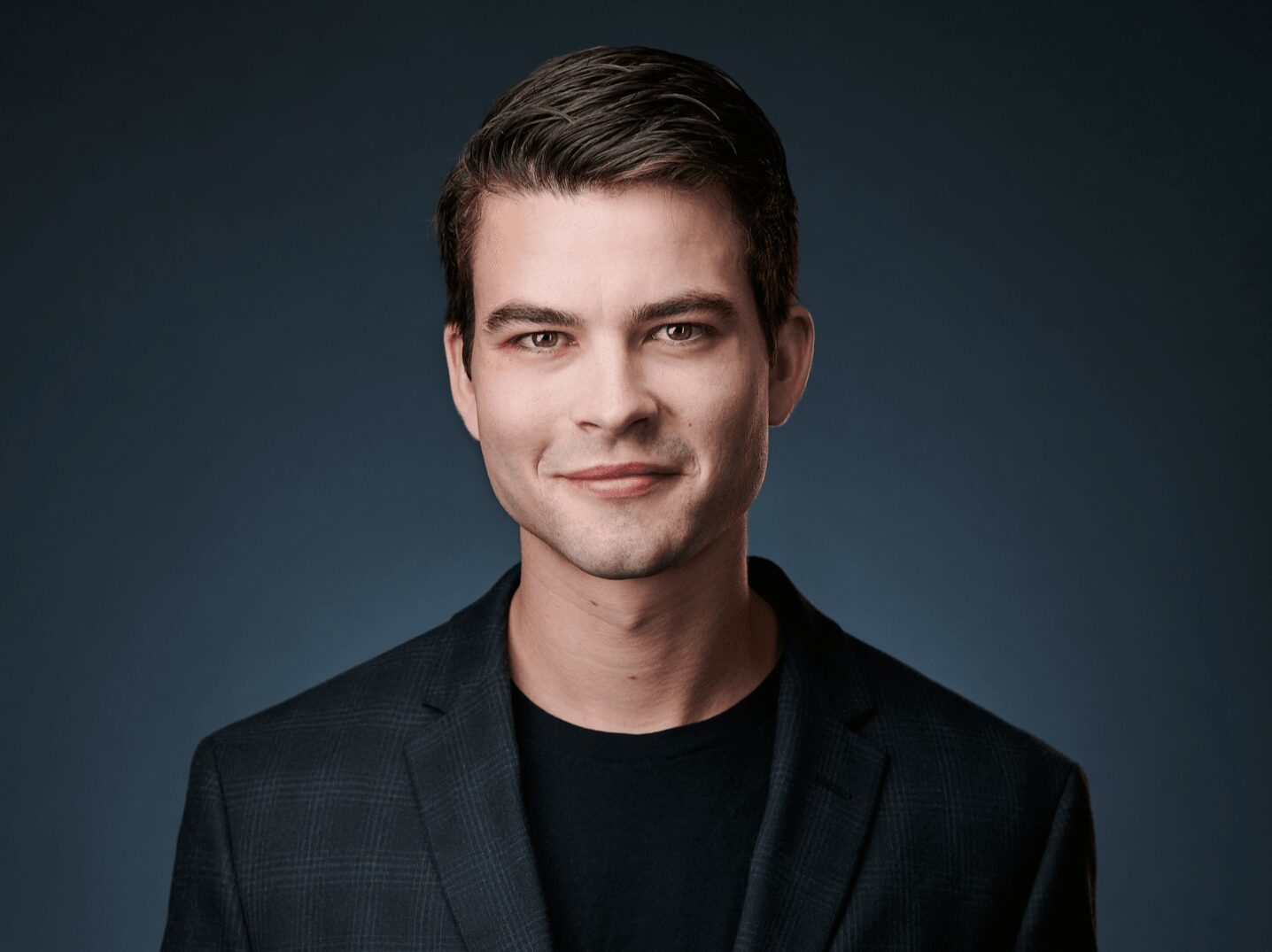 Young man with dark hair and a slight smile, wearing a black blazer against a dark background.