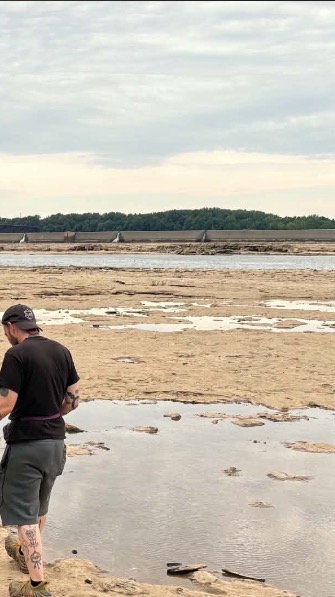 Person standing on a sandy beach near water, with a cloudy sky and distant land in the background.