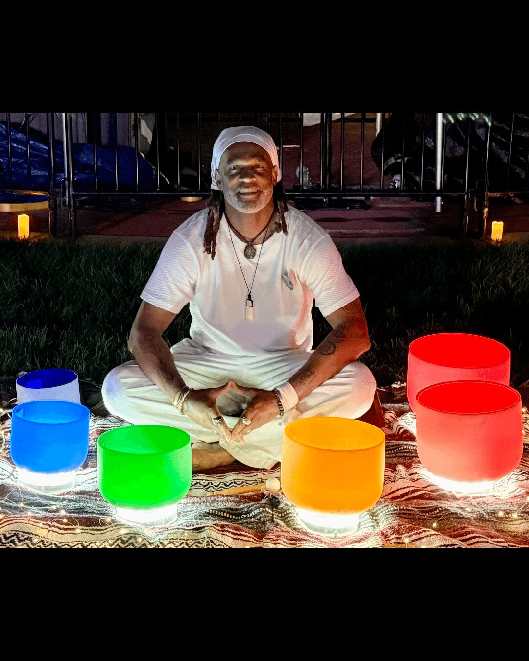 Person sitting cross-legged on a colorful mat with five illuminated bowls in front, outdoors at night.